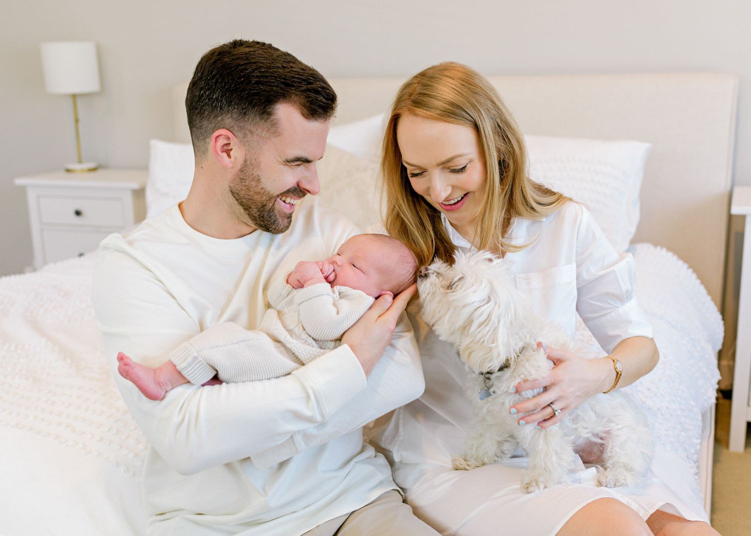 Couple in white clothing cuddles newborn baby while small white dog joins them on bed.