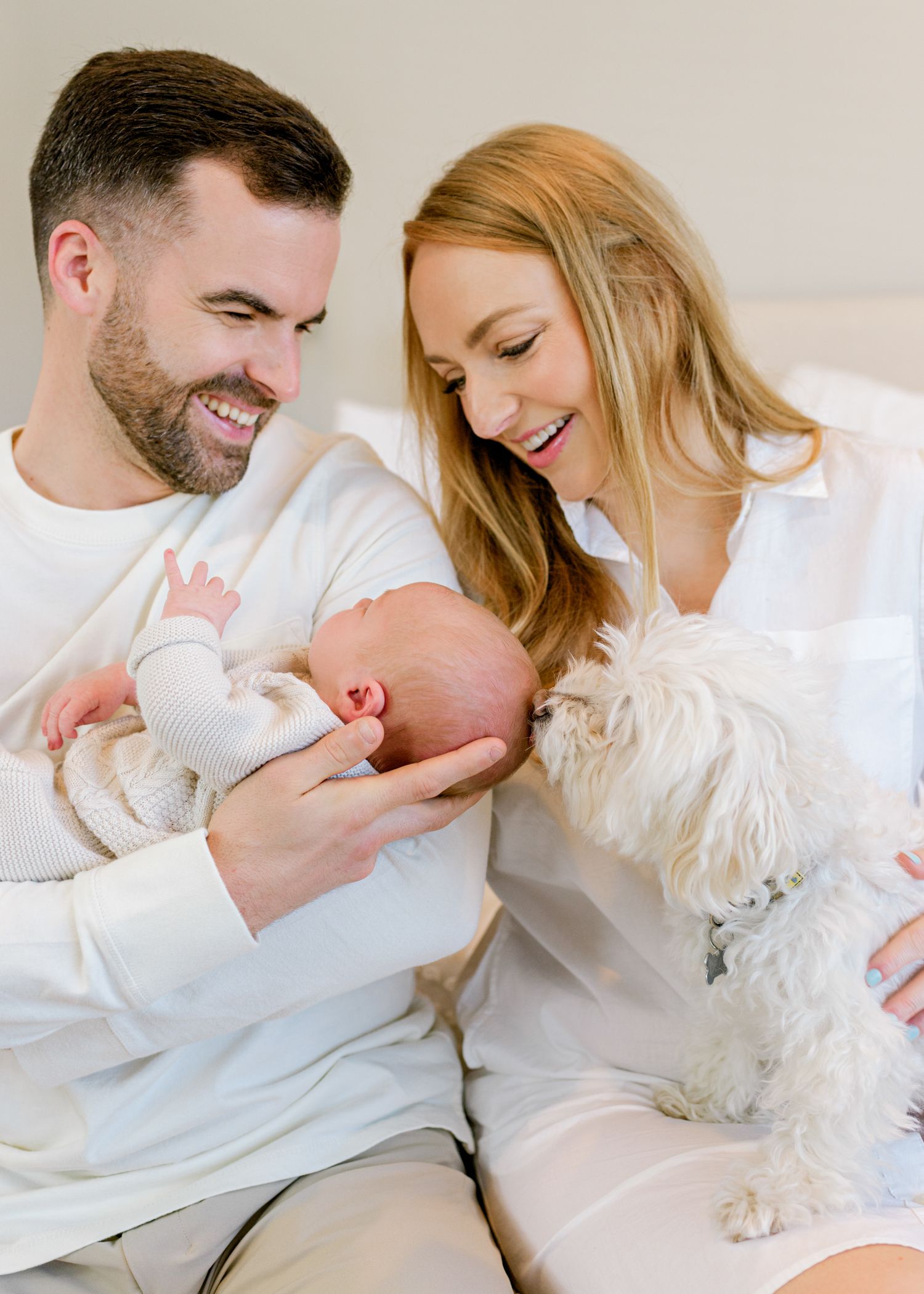 Family shares joyful moment with newborn baby and white dog on cozy bed.