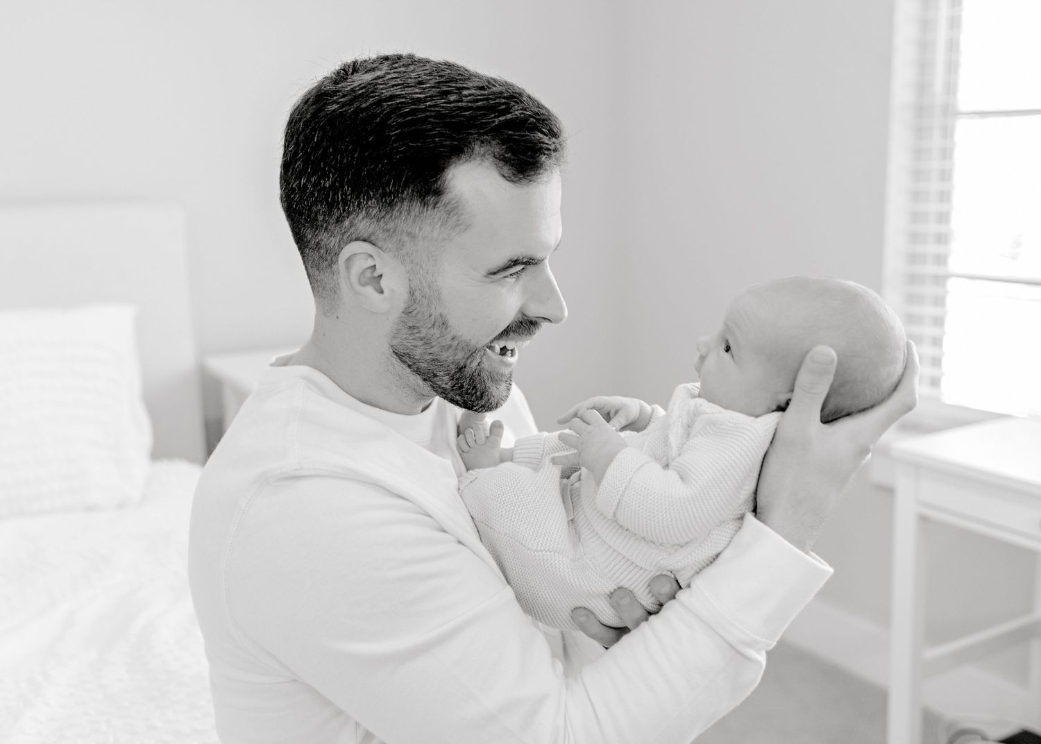 Black and white portrait of tender moment between parent and baby on white bed.