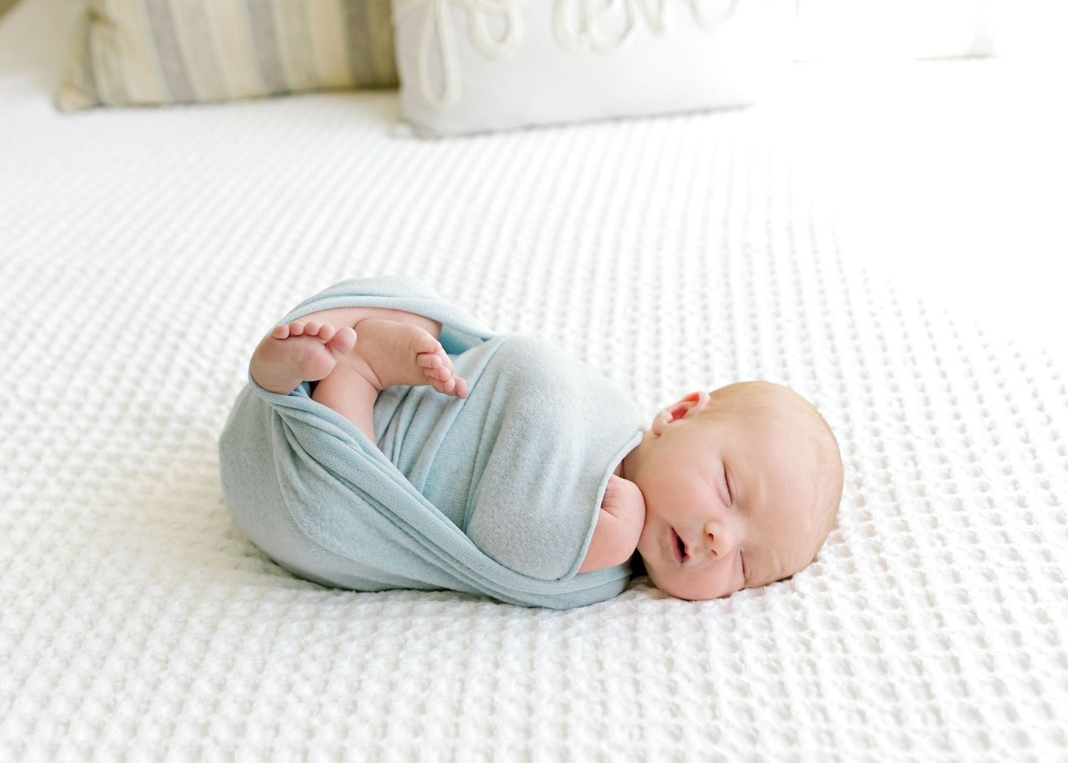 Sleeping newborn baby swaddled in light blue fabric on white textured bedding.