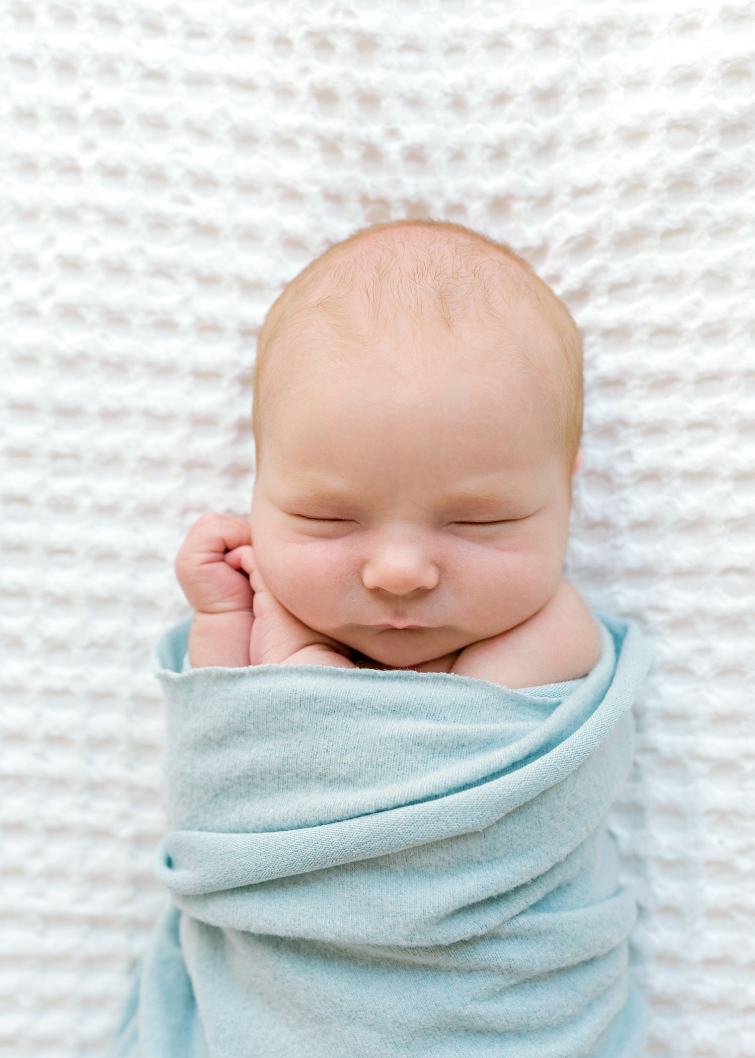 Peaceful sleeping newborn wrapped in soft blue swaddle against white textured background.