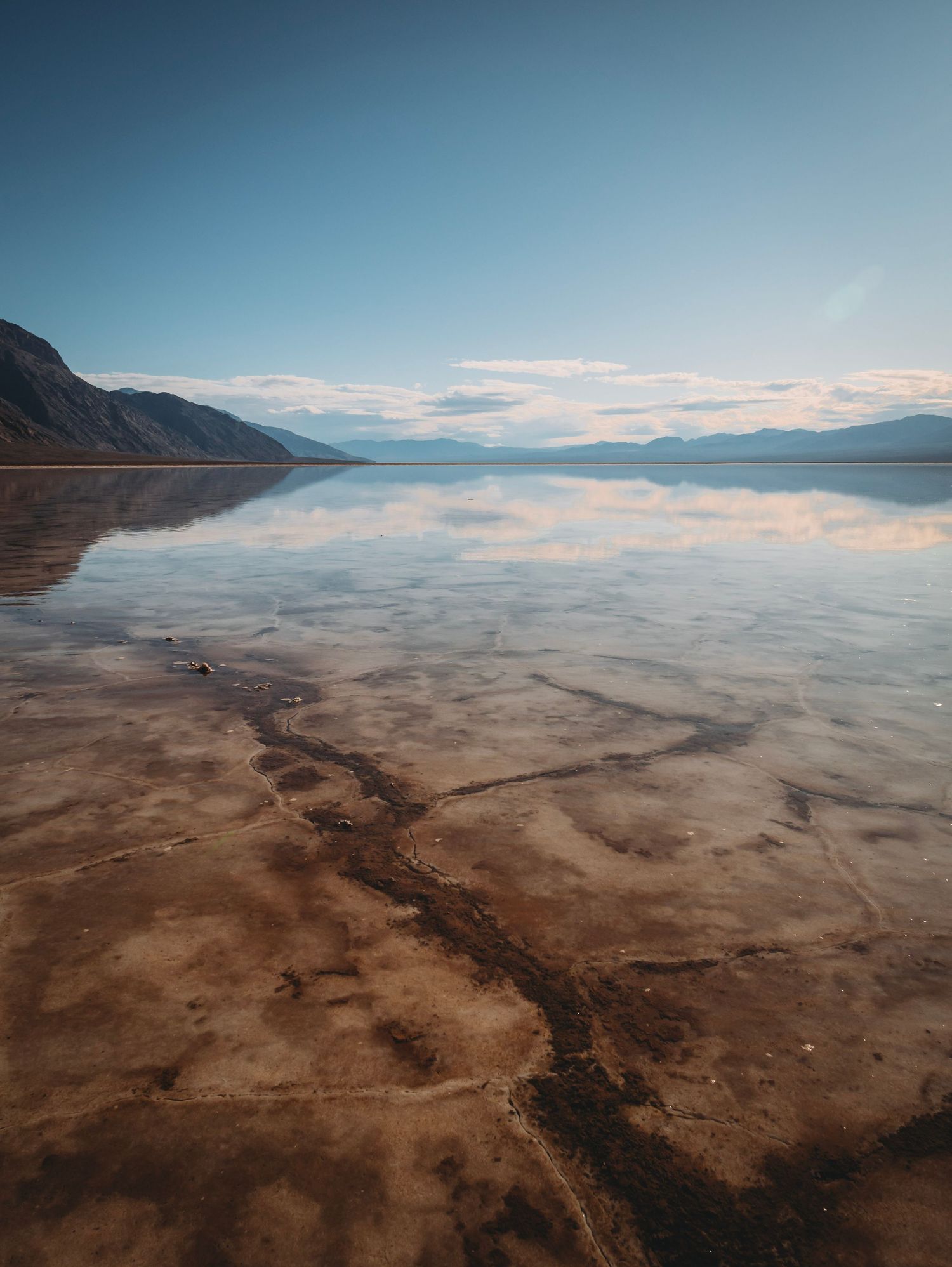 Calm lake surface mirrors mountain landscape and clouds with brown shoreline patterns.