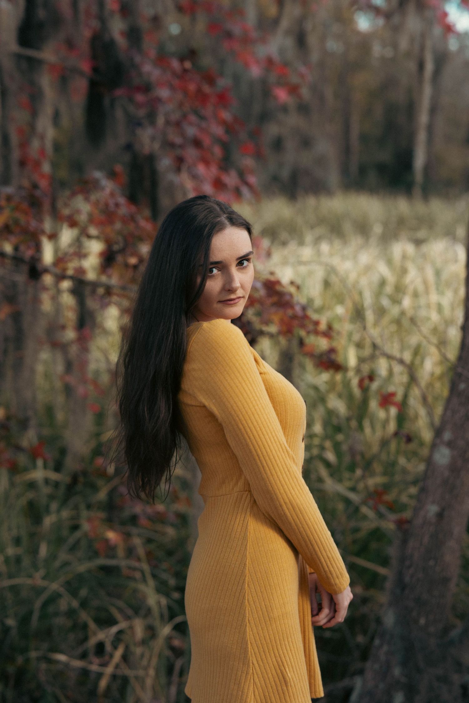 Woman in mustard yellow sweater dress poses against autumn foliage background.