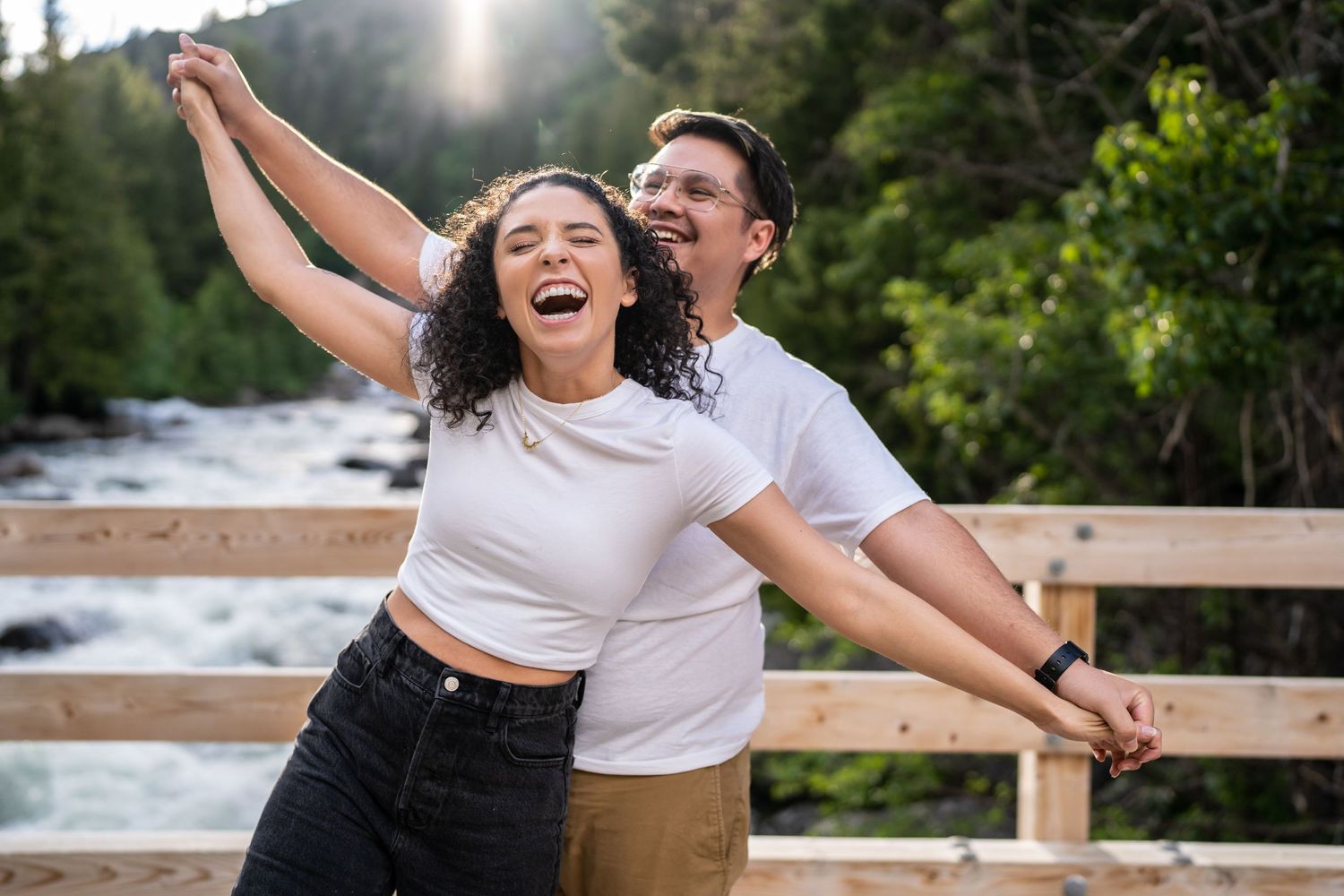 A joyful couple poses playfully on a wooden bridge with a rushing waterfall in the background.