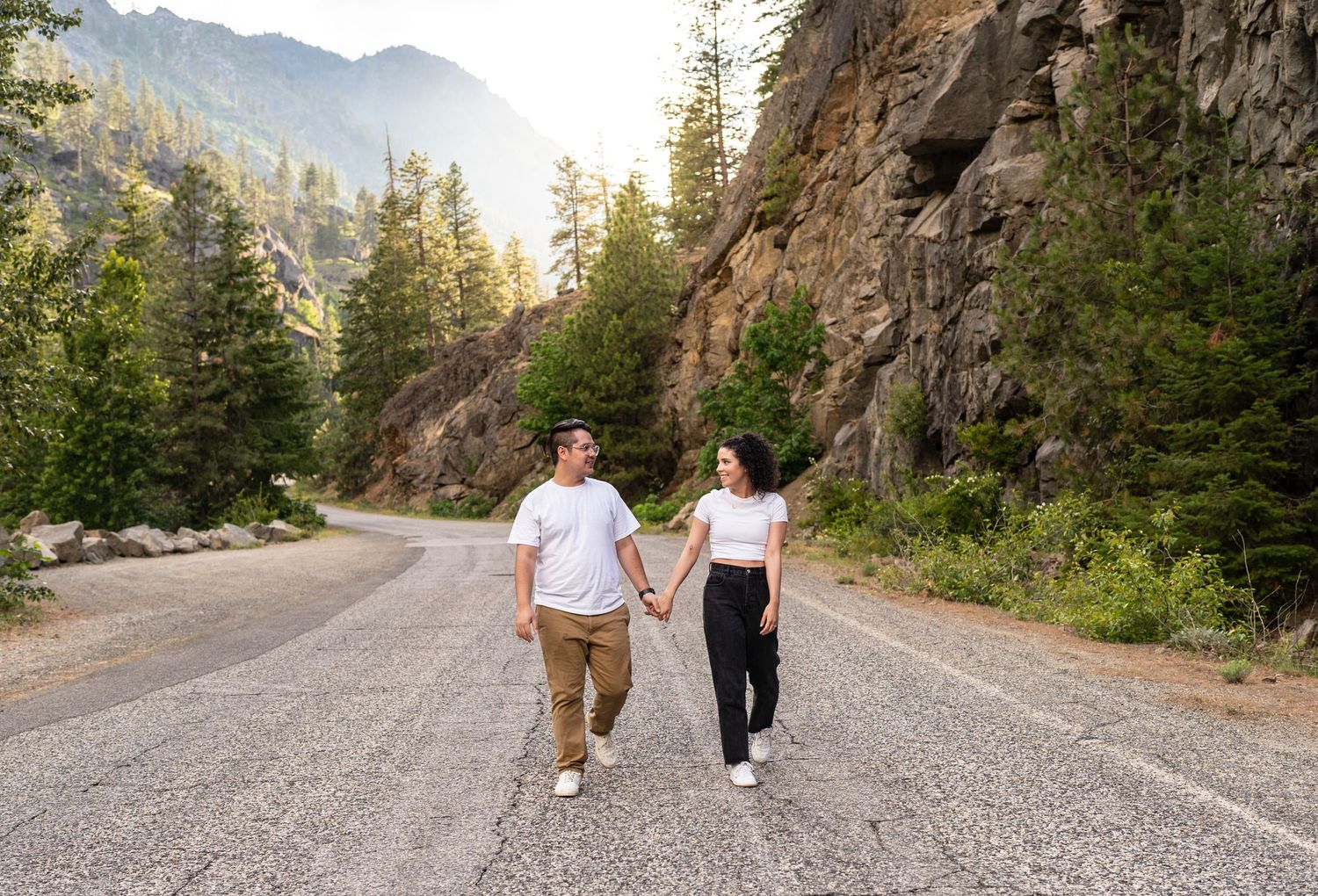 Two people walking together on a scenic mountain road surrounded by evergreen trees and rocky cliffs.