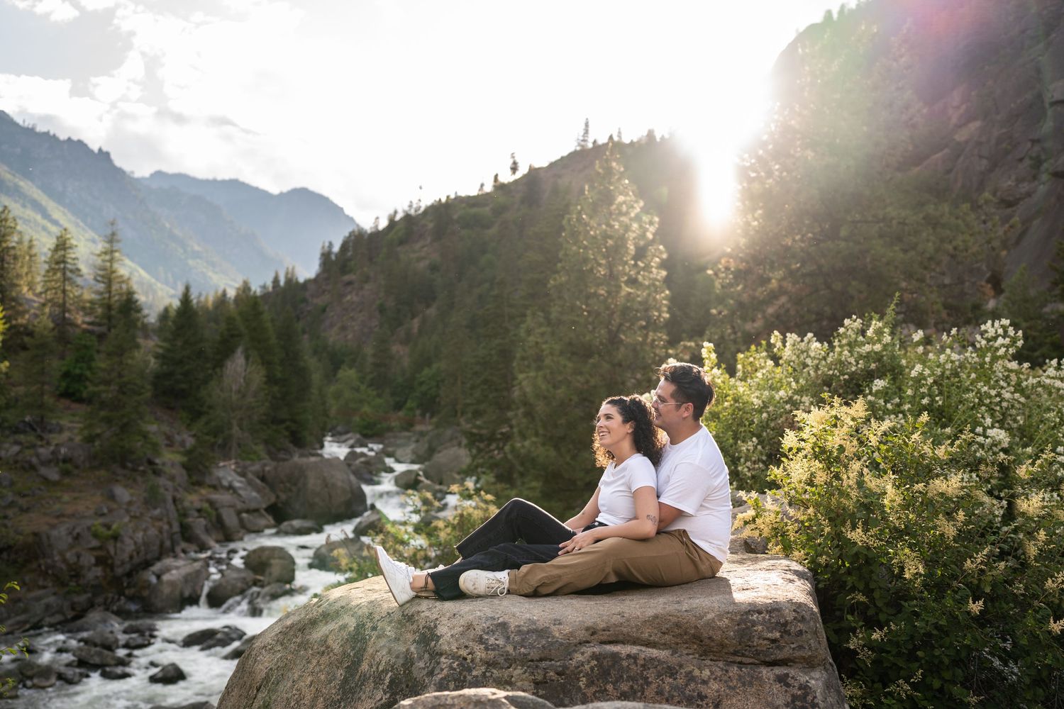 Couple relaxing on a rocky outcrop overlooking a mountain stream at sunset.