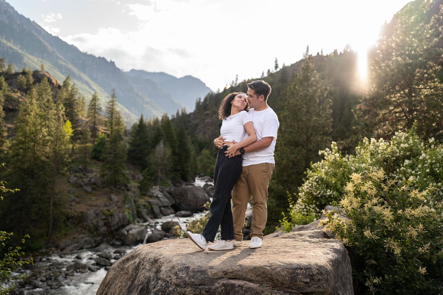 Two people embrace on a rocky overlook with a scenic mountain stream and forest backdrop.