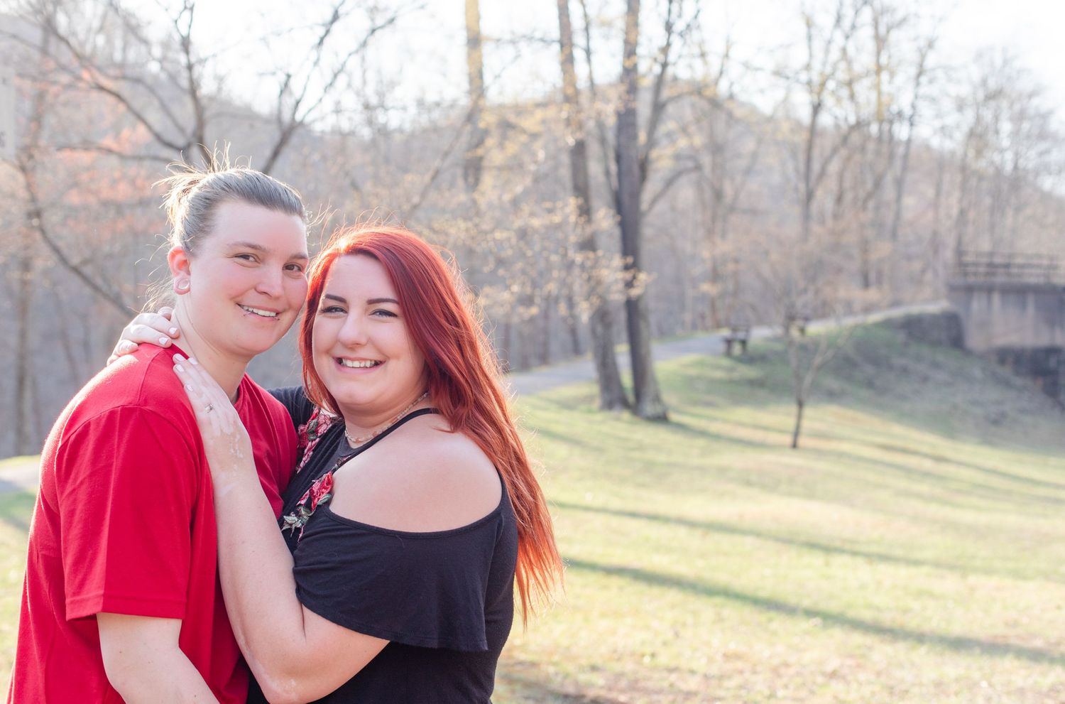 A couple poses together outdoors on a sunny day with trees and a bridge in the background.