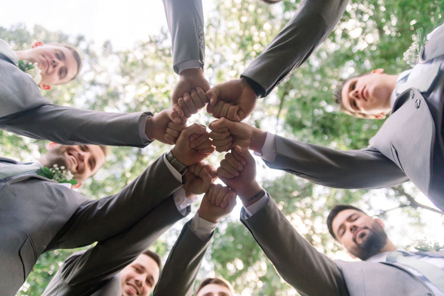 Groomsmen join hands in a circle formation under green trees.