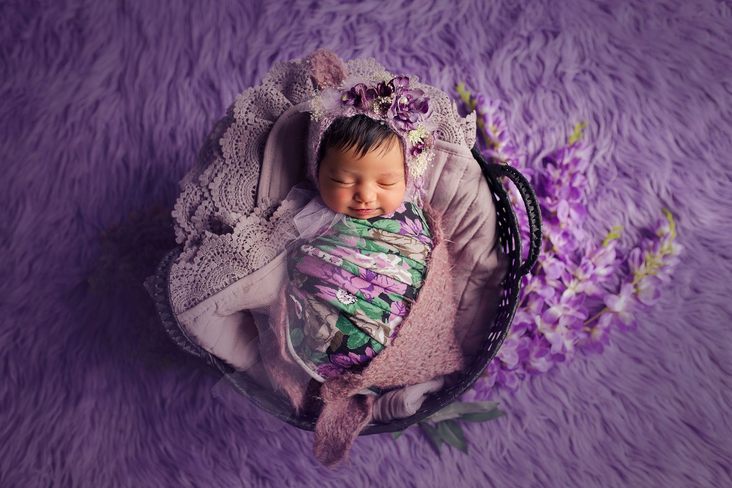 bebe sonriendo dentro de una canasta con flores escenario violeta y corona de flores