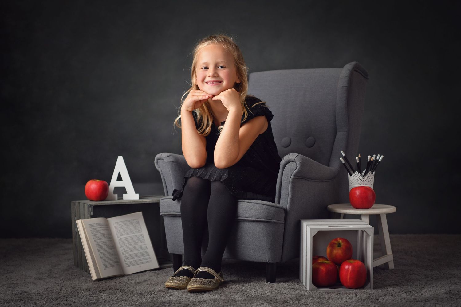 Studio portrait with grey armchair, red apples and books against dark background.
