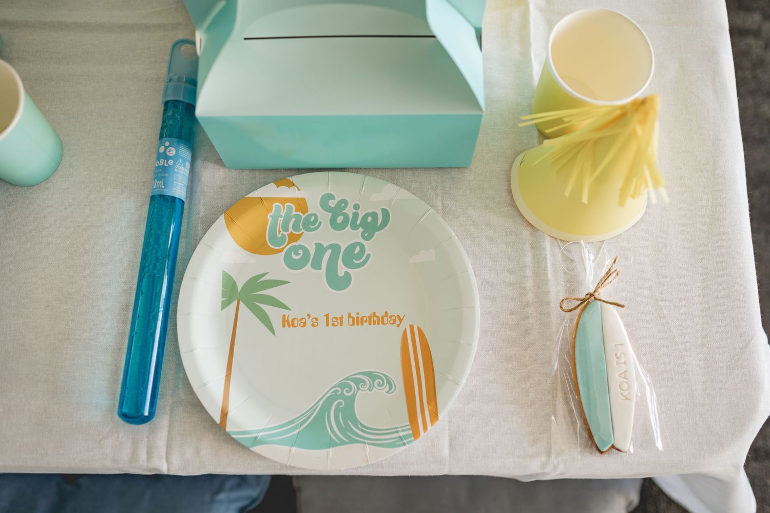 Place setting with wave-themed plate, party favor box, and decorative elements in mint and yellow.