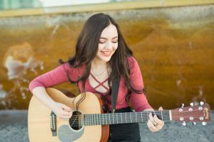 A person plays an acoustic guitar while wearing a burgundy top in an outdoor setting.