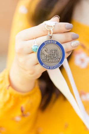 A hand wearing sparkly nail polish holds up a blue and silver high school medallion against a yellow background.
