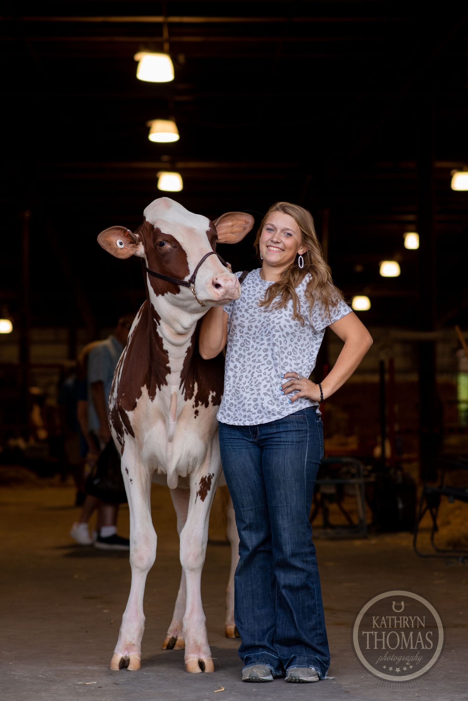 A tall brown and white dairy cow stands with its handler in a dimly lit barn.