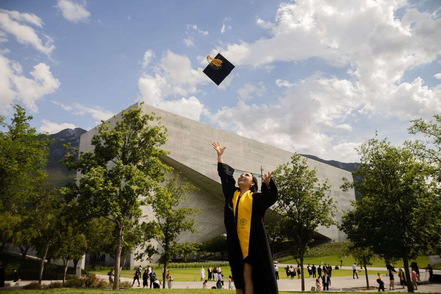 Graduate in black and gold regalia throwing cap into sky near modern university building.