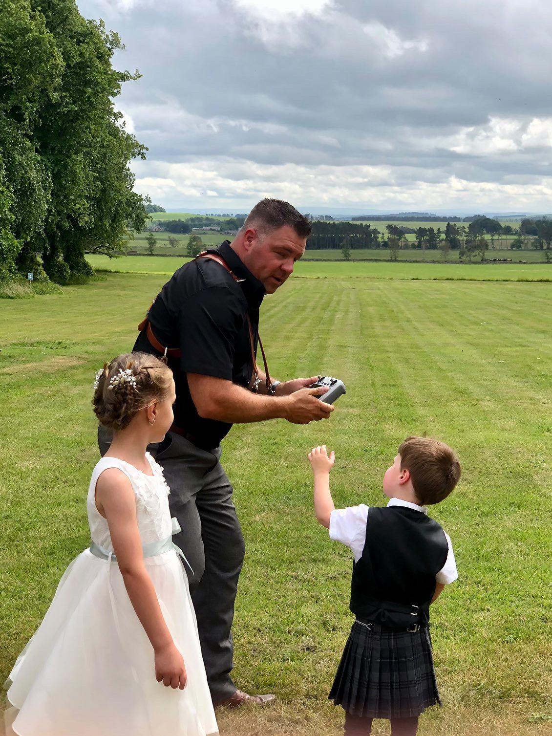 Wedding photographer showing photos to children in formal attire on grassy hillside.