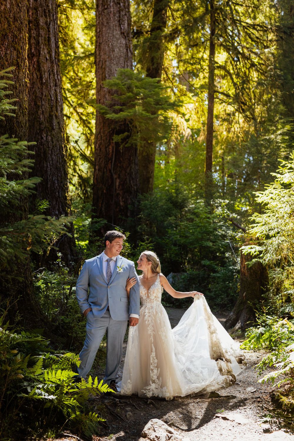 Bride in lace gown and groom in grey suit walk a sunlit forest path among tall trees.