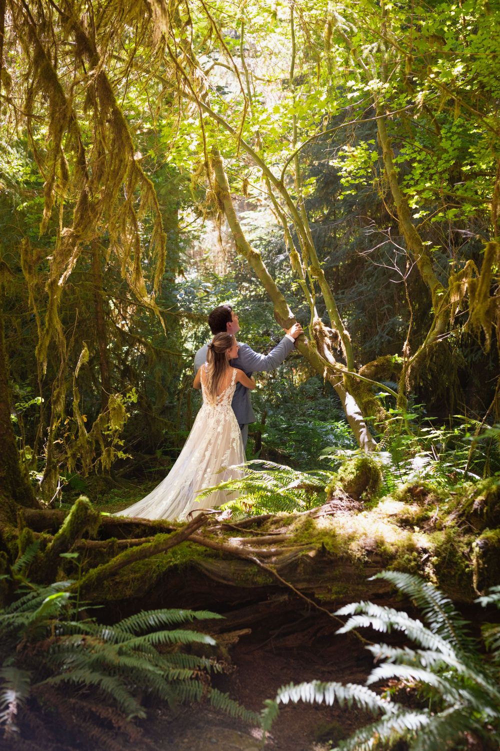 Couple explores a magical moss-draped forest, groom pointing toward sunlit canopy.