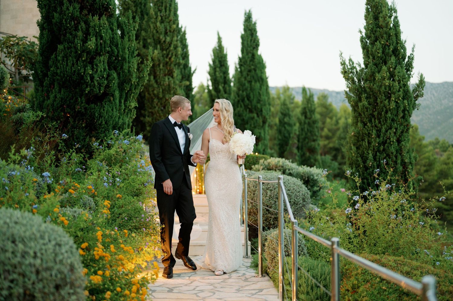 A newlywed couple walks down garden steps lined with cypress trees and yellow flowers.