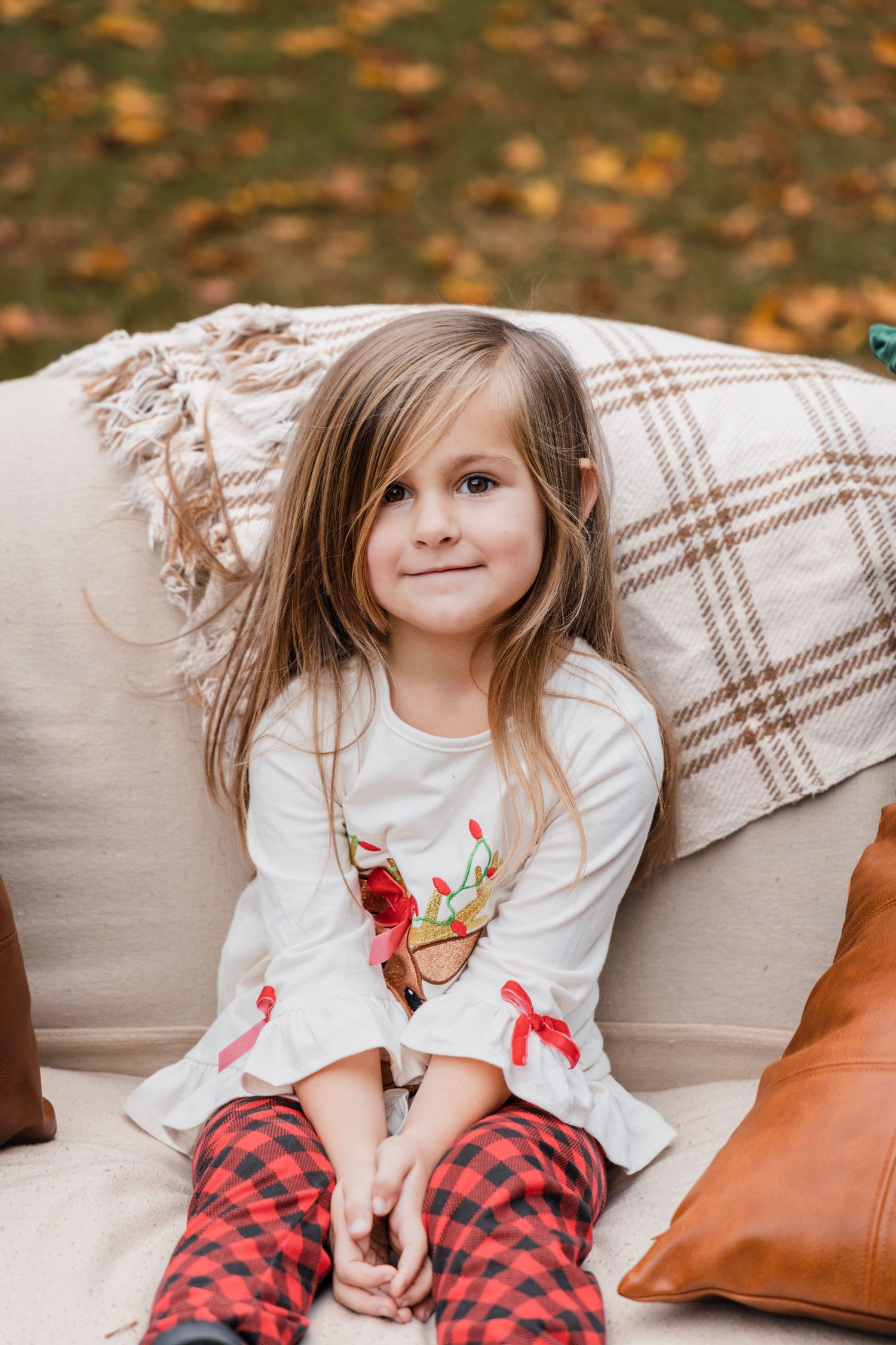 Child in plaid blanket and white shirt sits on couch with autumn decor and orange pillows.