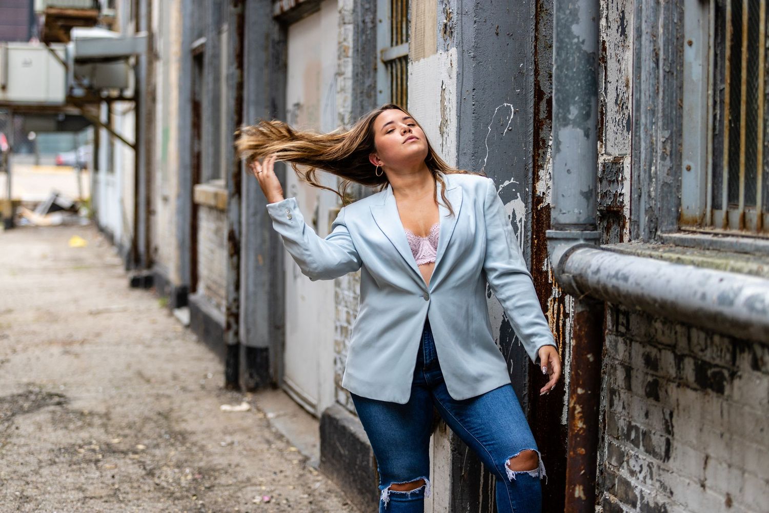 Confident woman in blue blazer and ripped jeans poses against weathered urban building wall.