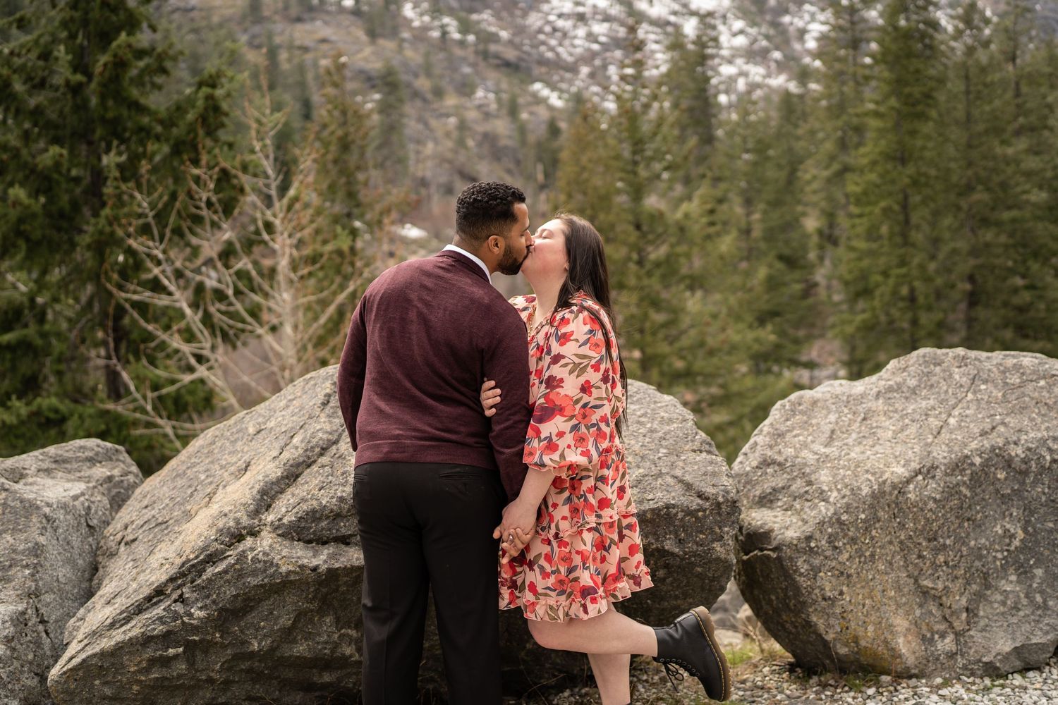 A couple embracing near large boulders with evergreen trees and mountains in the background.