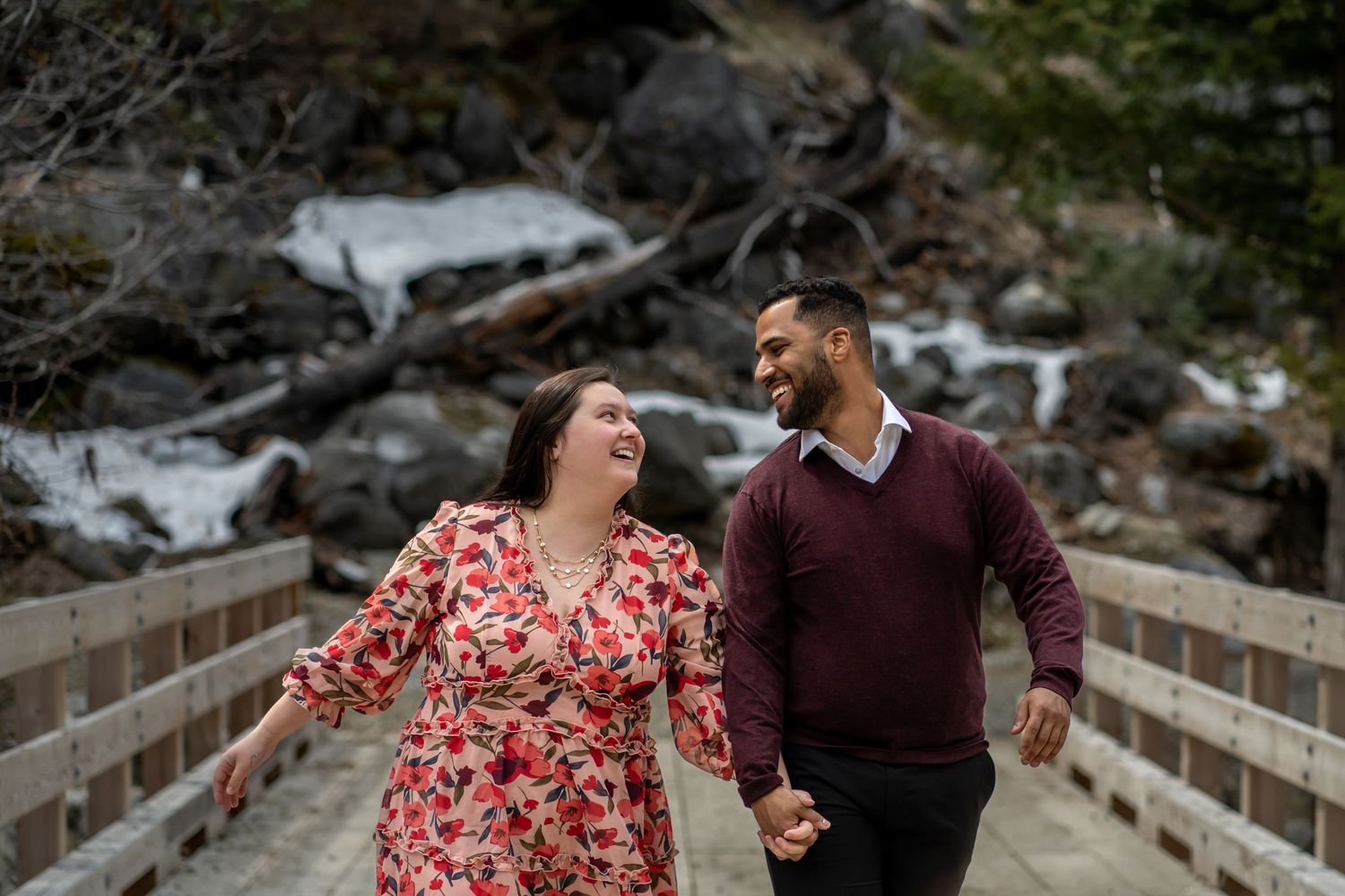 A couple laughing together on a wooden bridge surrounded by snowy mountain scenery.