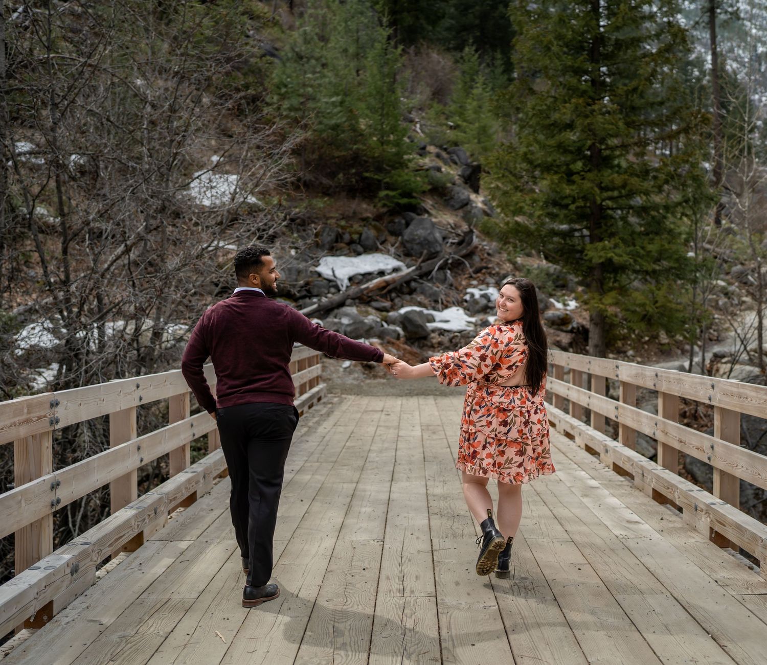 A couple dancing and holding hands on a wooden bridge surrounded by evergreen trees and mountains.