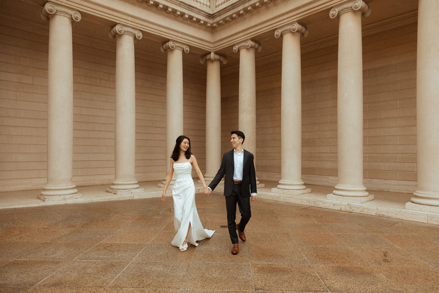 Elegant couple walking hand in hand through classical colonnade with white pillars.