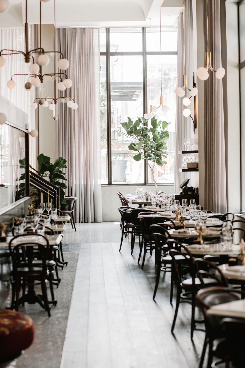 Elegant restaurant interior with black bistro chairs, white globe pendant lights, and tall windows with draped curtains.