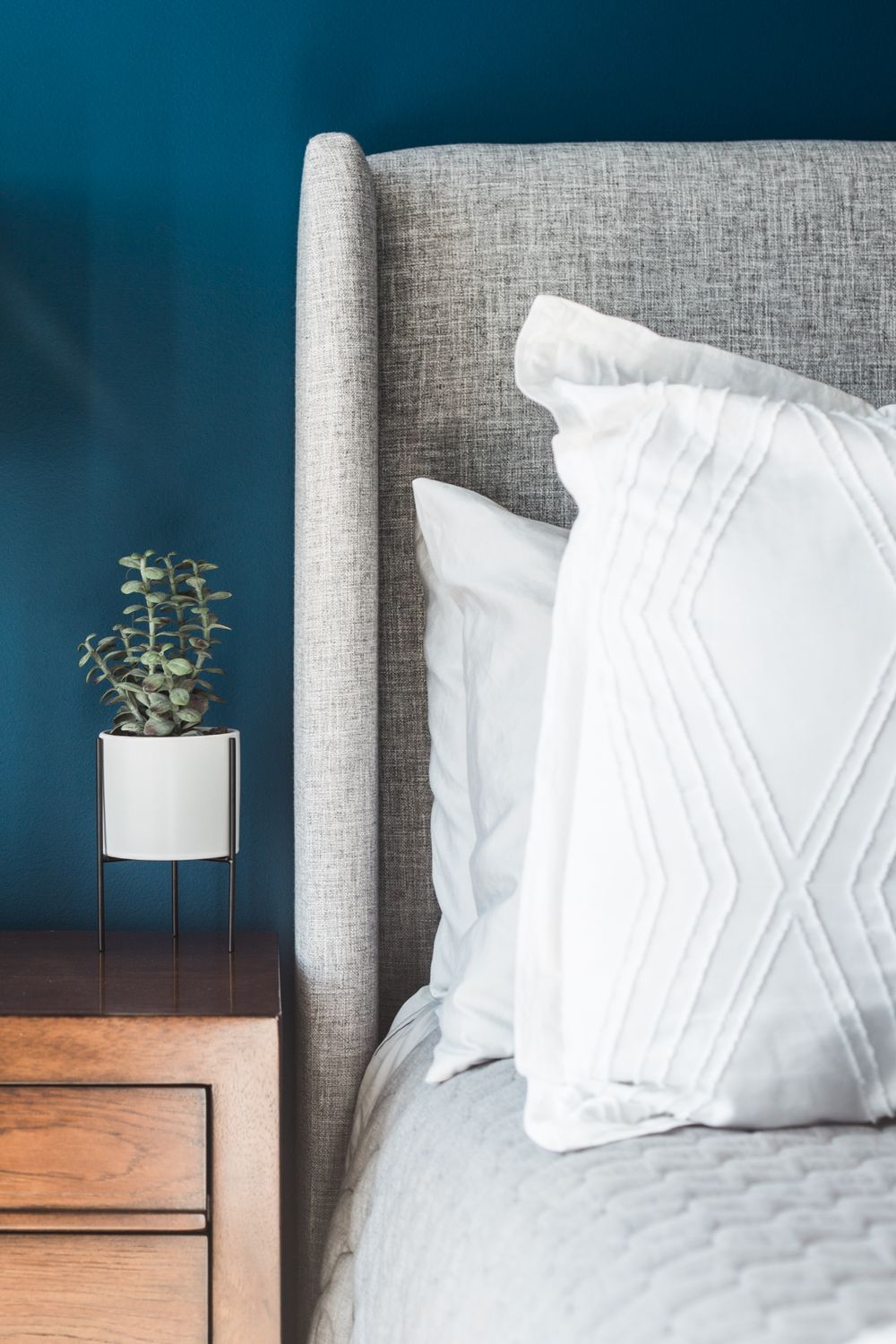 Gray upholstered bed with white pillows against teal wall, wooden nightstand with white potted plant.