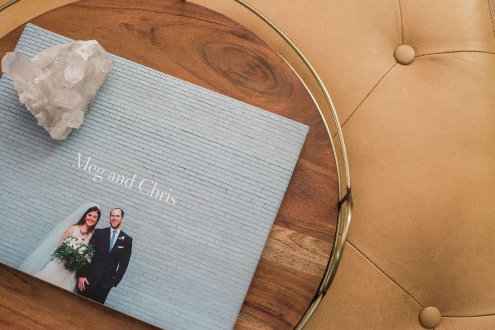 Wedding photo album for Meg and Chris resting on a wooden ottoman beside a gold leather chair with a quartz crystal.