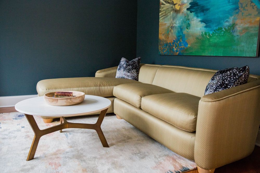 Gold leather sectional sofa with blue accent pillows beside a round marble coffee table in a teal-walled living room.
