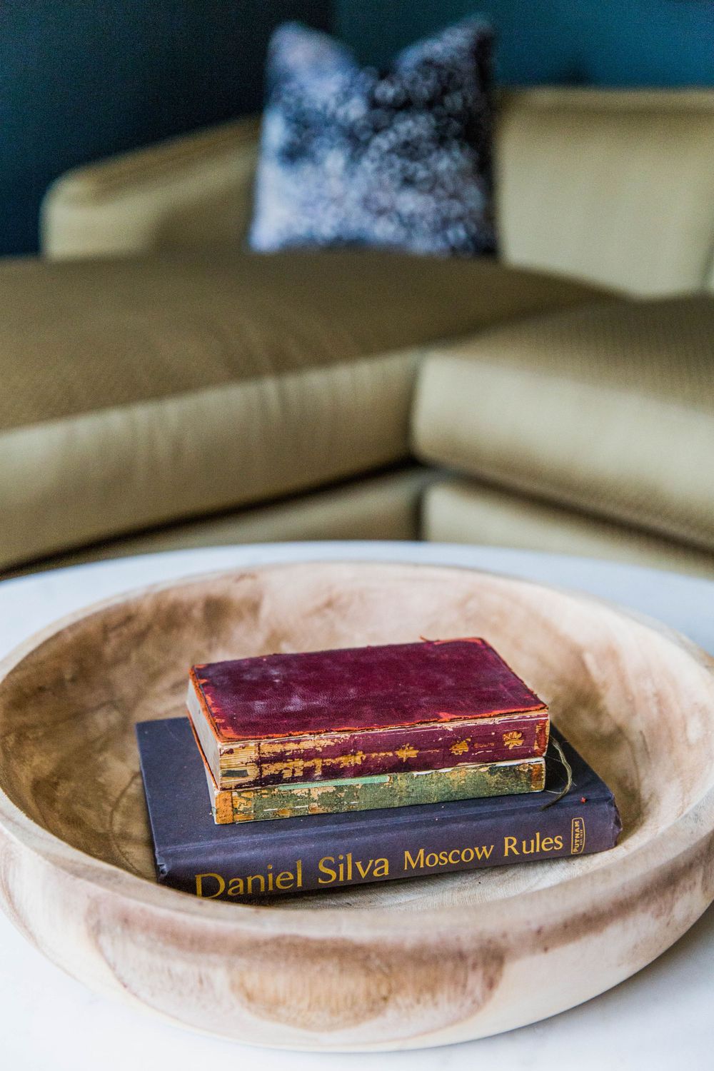 Vintage books including Daniel Silva Moscow Rules displayed in a decorative wooden bowl on a white coffee table.