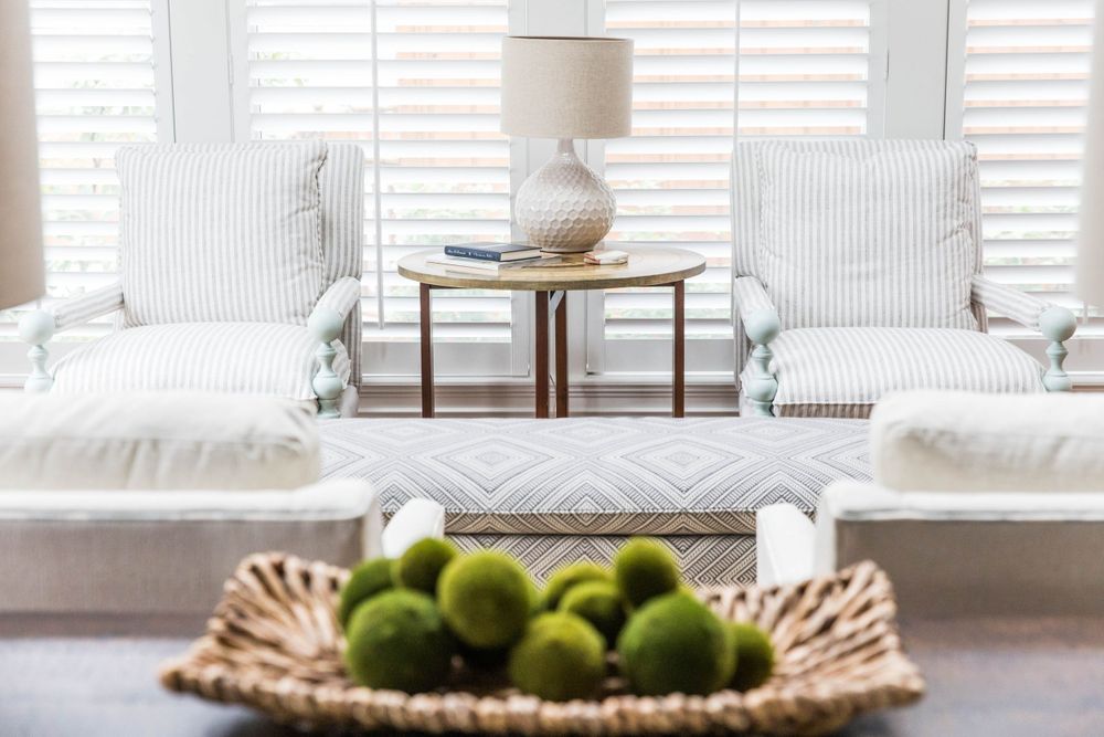 Bright living room with white striped armchairs, a woven lamp, and a basket of green moss balls on a dark table.