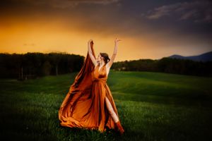 Figure in flowing orange dress dances in a meadow against dramatic sunset sky.