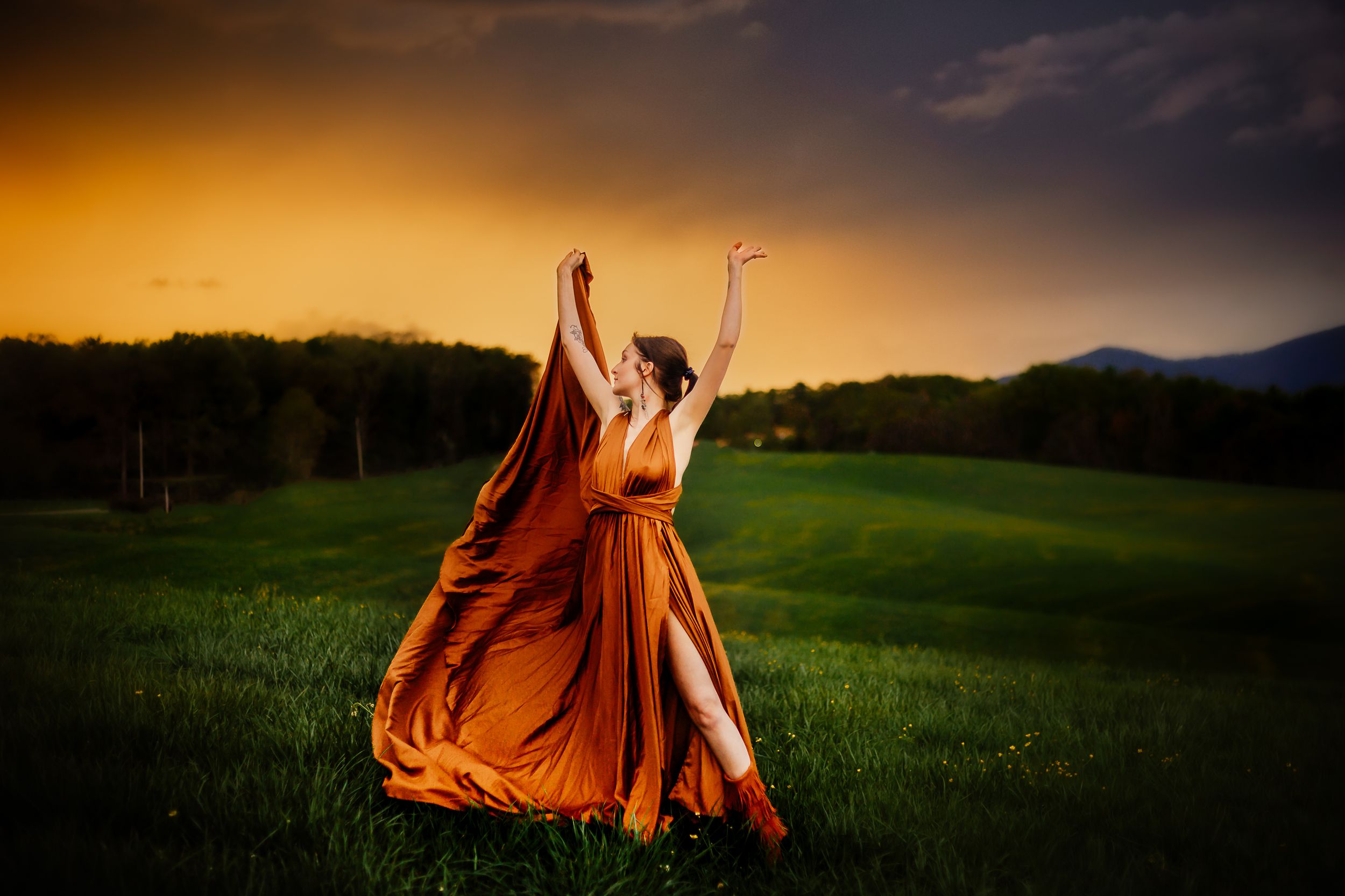 Figure in flowing orange dress dances in a meadow against dramatic sunset sky.