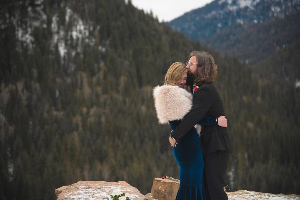 bride in a blue velvet gown with fur stole kissing groom on a cliff in the winter