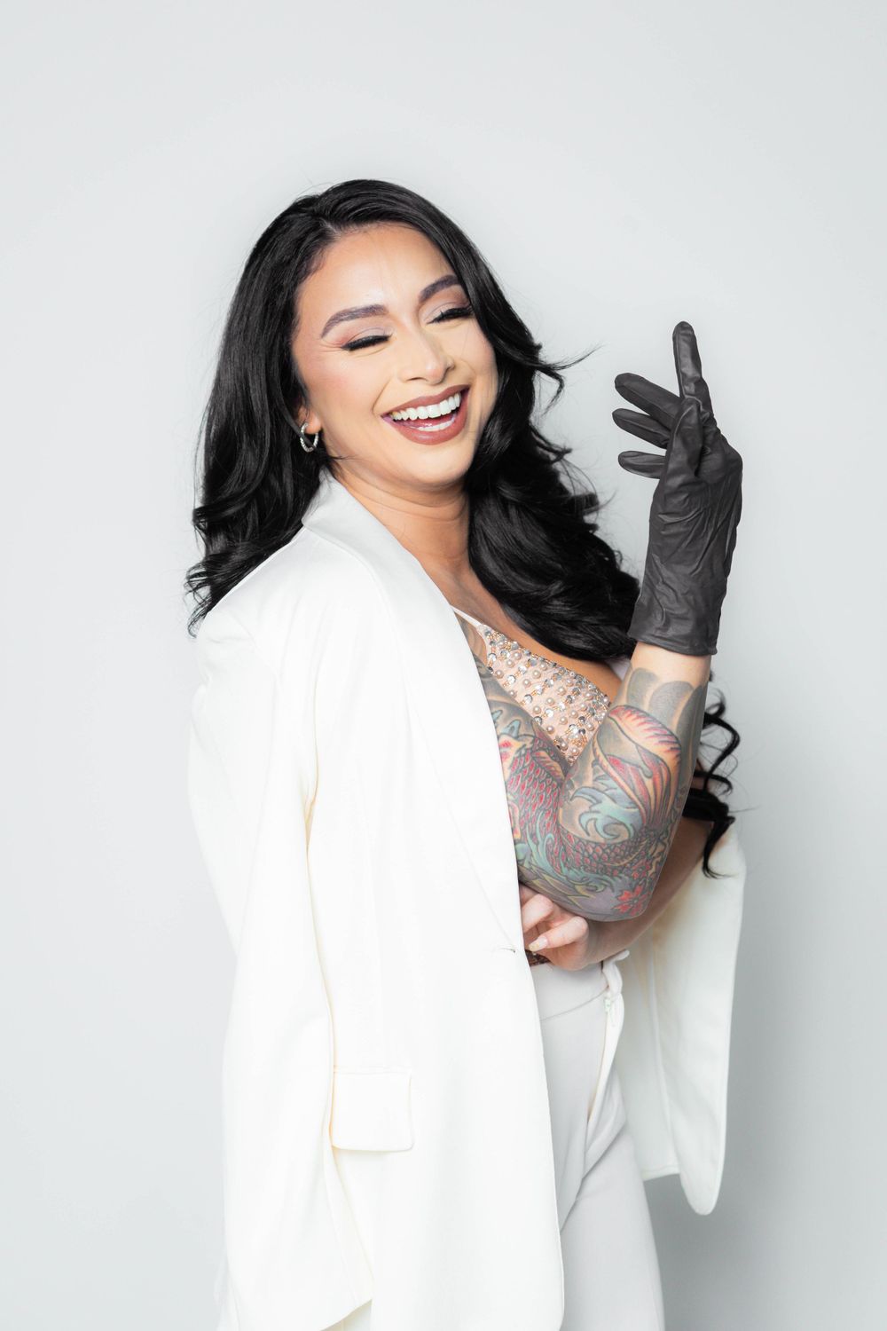 Laughing tattooed woman in white blazer wearing black glove on gray background.