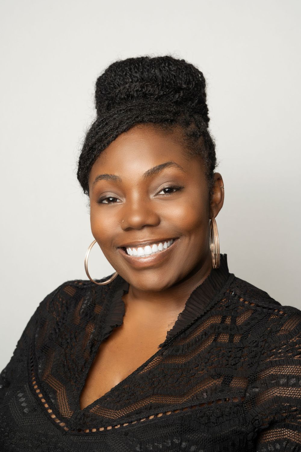 Smiling Black woman with updo hairstyle wearing hoop earrings and black lace top.