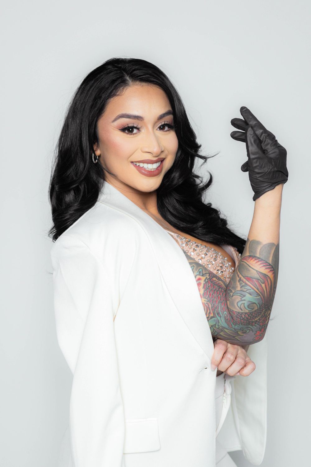 Tattooed woman in white blazer and black glove posing against light gray background.