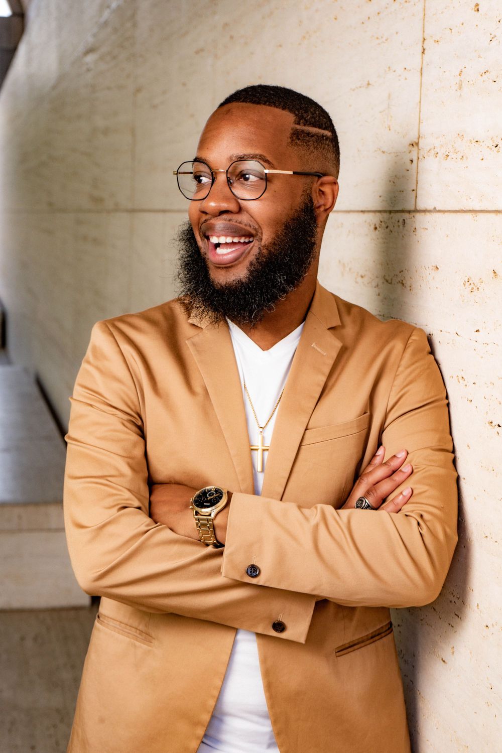 Stylish Black man with beard and glasses in tan blazer smiling with arms crossed.