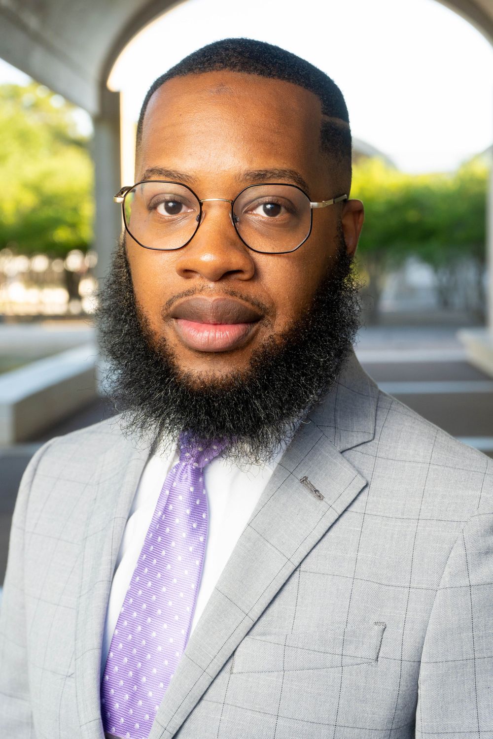 Professional Black man with beard and glasses wearing gray suit and purple tie outdoors.