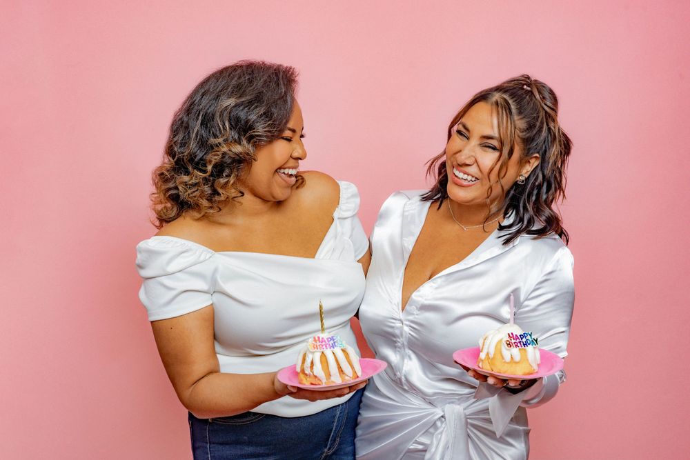 Two laughing women in white tops hold birthday cakes on a pink background.