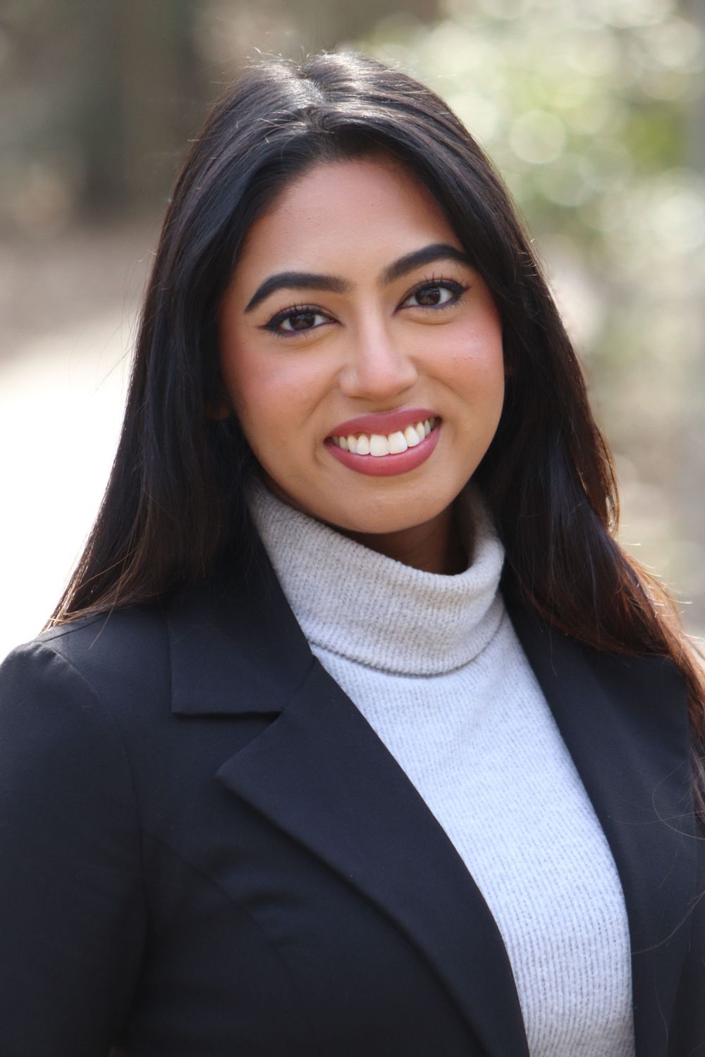 Professional headshot of a young woman in a black blazer and turtleneck sweater smiling outdoors.