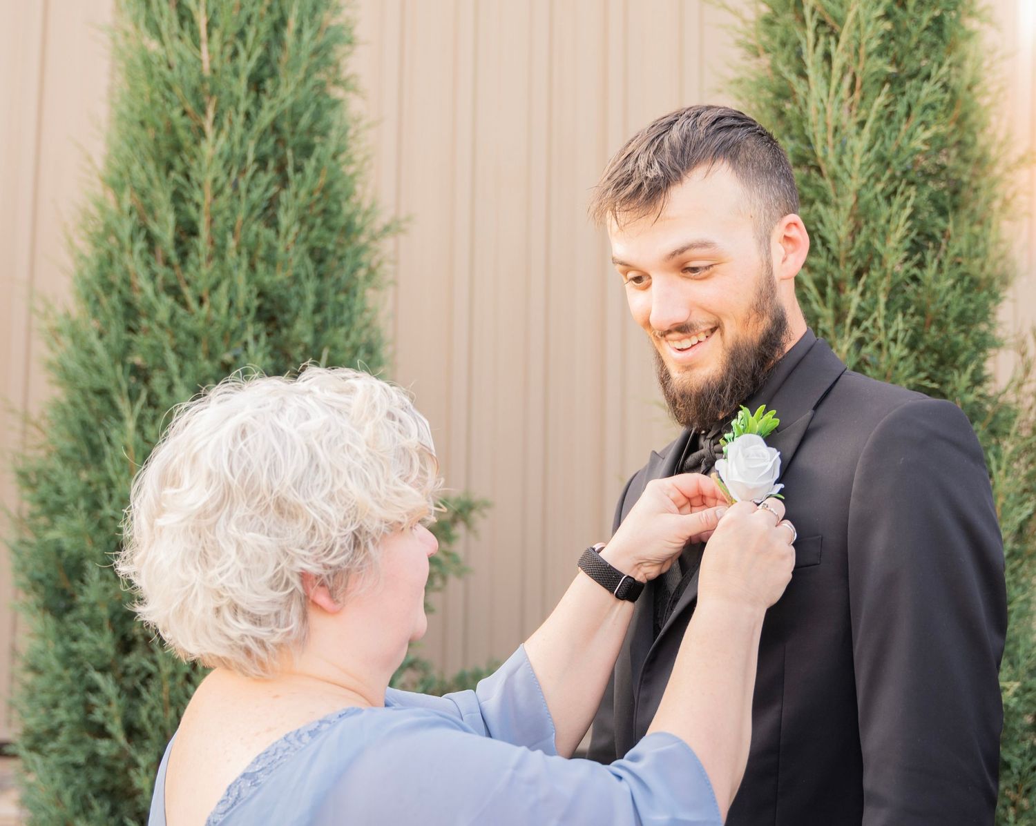 Older woman pinning a succulent boutonniere onto a smiling bearded groom in a black suit.
