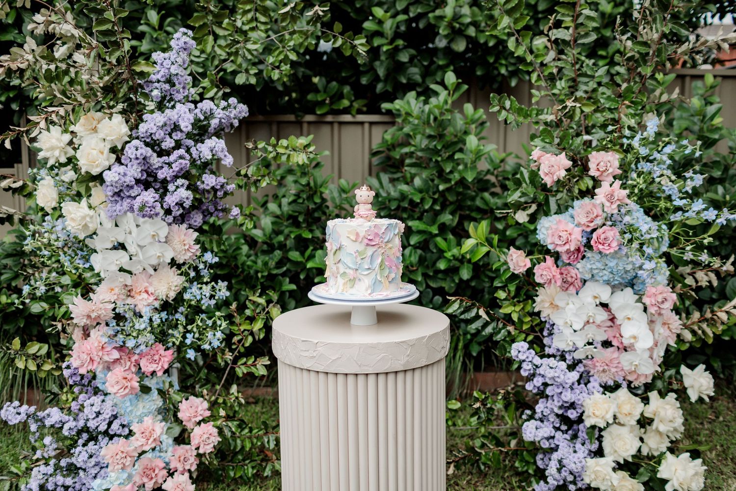 White cake displayed on ribbed pedestal between two floral arches in garden setting.
