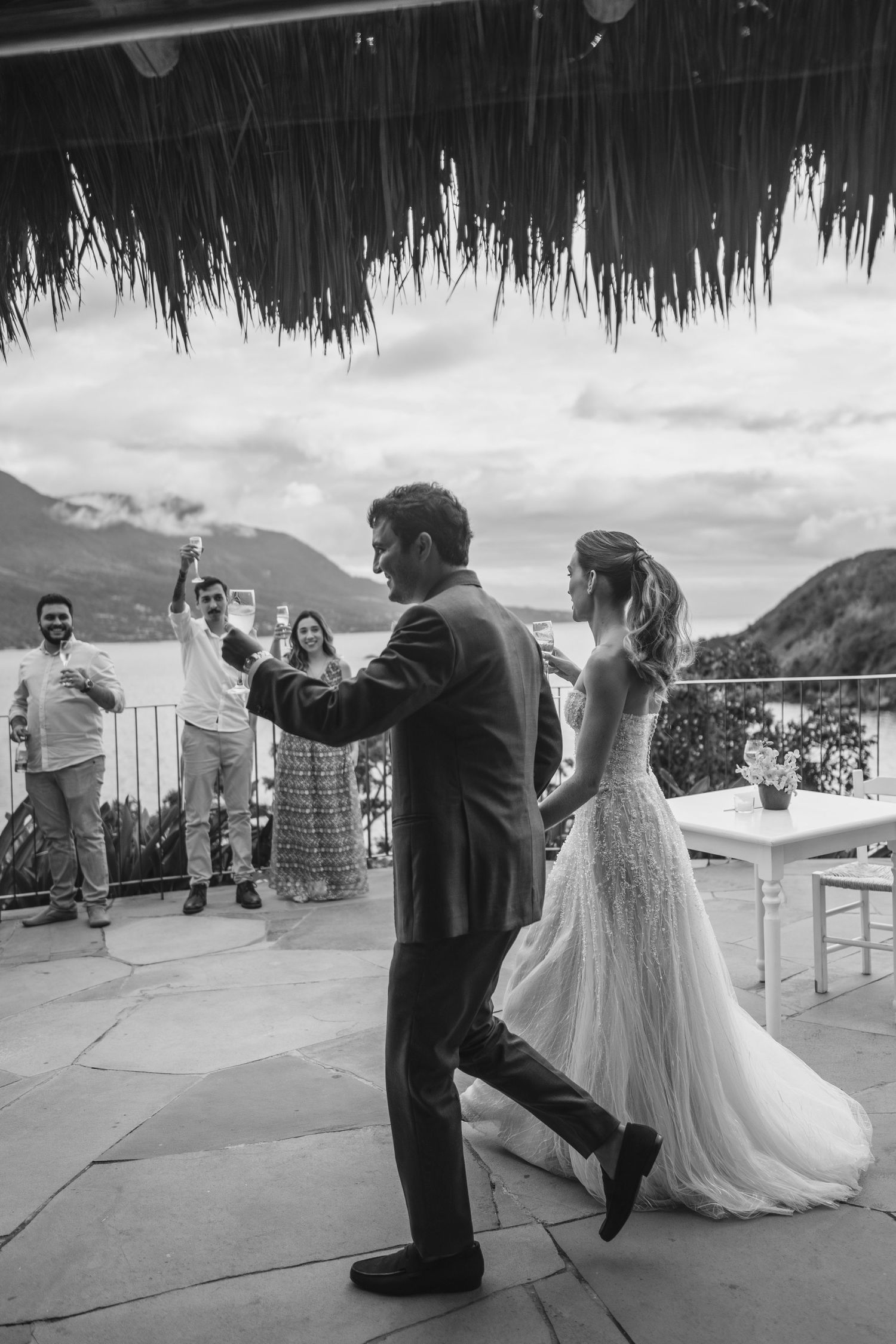 Black and white photo of wedding celebration dancing under thatched roof with lake view.