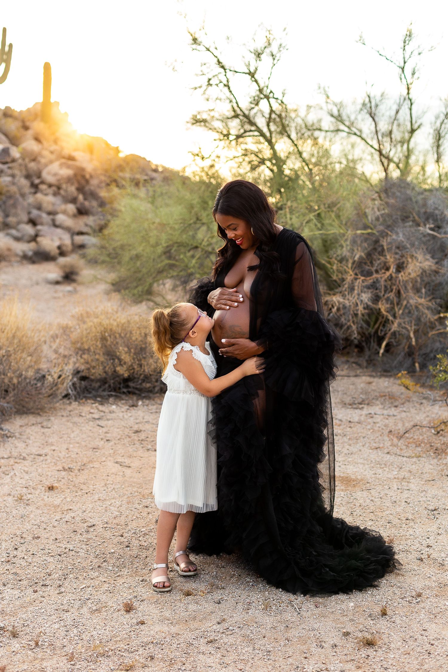Sunset maternity photo with young girl touching pregnant belly in desert setting.